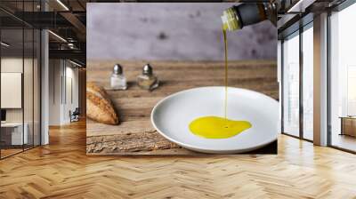 Extra virgin olive oil pouring from a bottle to a round white plate, on a rustic wooden table, with bread, salt and pepper. Wall mural