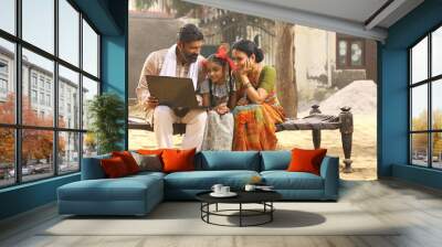 Happy Indian Rural family of Mother, father, daughter sitting on cot outside their home in front yard using Laptop. traditional wear sari kurta pajama Wall mural