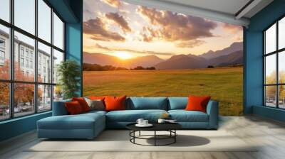 the sun is setting casting a warm afterglow over the grassy field with mountains in the background cumulus clouds dot the sky creating a picturesque natural landscape Wall mural