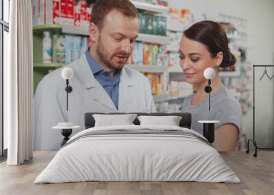 Attractive young woman talking to the chemist, shopping for medicament at the drugstore. Mature male pharmacist helping his female client Wall mural