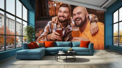 Happy bearded man enjoying drinking beer with his best friend. Male friends hugging, holding up their beer glasses, celebrating at the bar Wall mural