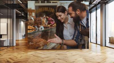 Young beautiful woman buying delciious freshly baked pastry at local bakery. Professional baker pointing at the display, talking to his customer. Bearded mature man working at his cafe, selling desser Wall mural