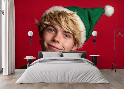 A young man dressed as a cheerful elf, with tousled blonde hair and mischievous green eyes, on a vibrant red background, adding to the holiday spirit 1 Wall mural