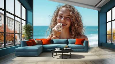 Teenage girl with curly hair, wearing a summer dress, eating an ice cream cone with the ocean in the background 3 Wall mural