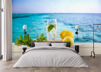 Glass of still mineral water with ice and fresh mint and lemons on wooden board table and tropical blue seascape background. Wall mural