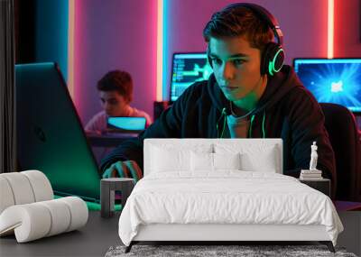 A Boy wearing headphones using a laptop with neon lights glowing background slide view. A teenage gamer typing or on her notebook keyboard sitting at a desk. An engineering student is Coding. Wall mural