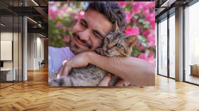 close up. Young man hugs a gray tabby cat with green eyes in the garden and smiles Wall mural