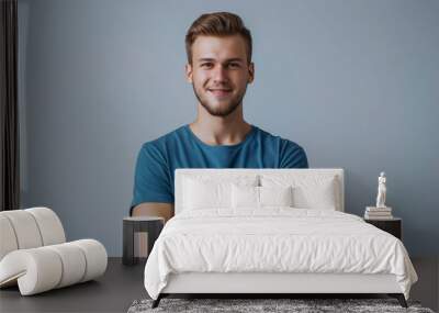 Indoor portrait of a young European Caucasian man isolated on a grey background, standing in a blue t-shirt with crossed arms, smiling and looking straight at the camera. Wall mural