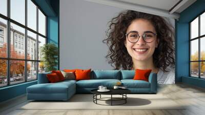 Portrait of a smiling young woman with curly hair and eyeglasses, set against a grey background. Wall mural