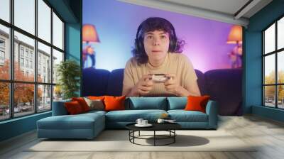 Portrait of young cute gamer woman sitting on sofa, playing online video game with wireless controller on console at home. Young gamer girl wearing headphone  Wall mural