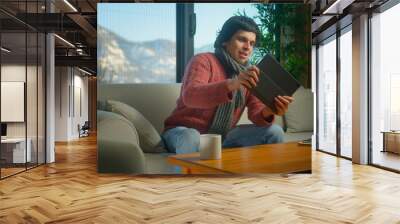 Young man wearing scarf playing a video game on a tablet computer while sitting on sofa in living room at home. Excited concentrated man engrossed in interesting game Wall mural