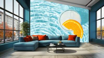 Glass of ice cubes with slice of fresh orange in rippled water of pool. Summer cold drink refresment concept. Pool party idea. Wall mural