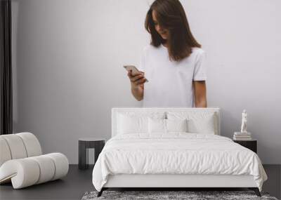 Photo of a smiling happy hipster girl wearing a casual white t-shirt using a mobile smartphone while standing on a white concrete wall background Wall mural