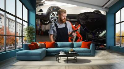 A male mechanic stands with a tablet and looks at a repair application against the background of a professional service station Wall mural