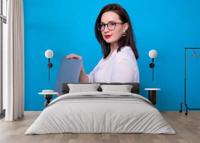 Portrait of a young brunette woman with a laptop on a blue background Wall mural