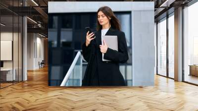 Powerful woman multitasking with her phone and laptop while standing outside among towering office buildings Wall mural