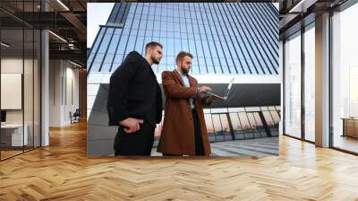 Two young guys discuss their business project and look at information on a laptop against the background of an office building Wall mural