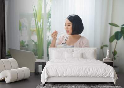 Beautiful pregnant business woman is eating yogurt and smiling while sitting at her working place at home Wall mural