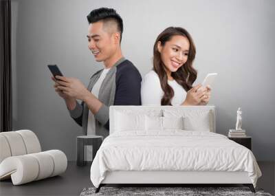 Beautiful young asian couple holding mobile phones and standing back to back against grey background Wall mural