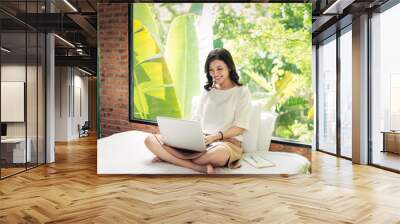 Beautiful young smiling woman working on laptop while sitting on bed at home near big window. Wall mural