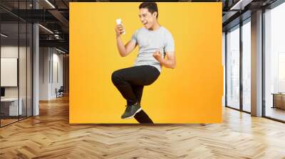 Cheerful young Asian man dressed in t-shirt jumping over orange background make winner gesture. Wall mural