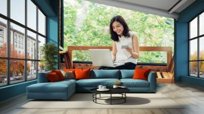 Confident young asian woman in smart casual wear shopping online while sitting near window in creative office or cafe Wall mural