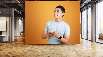 Portrait of a happy young asian man using mobile phone at studio on orange background. Wall mural