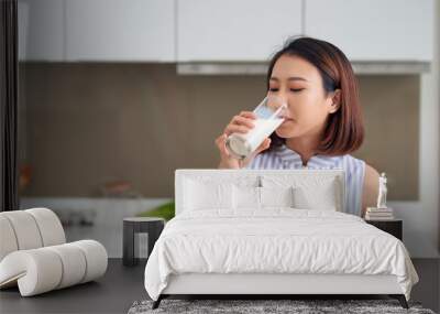 Portrait of young Asian woman drinking milk while standing in the kitchen. Wall mural