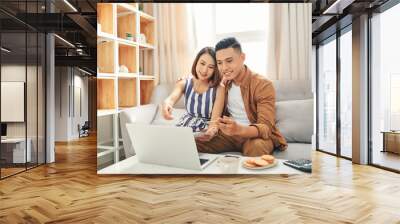 Smiling young couple sit on couch using laptop taking care of utility bills and house, read paperwork Wall mural