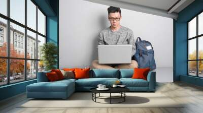 Young asian man in casual clothes is using a laptop, smiling while sitting on the floor Wall mural