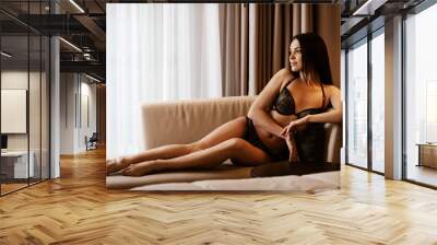 Beauty of woman body and lingerie concept. Beautiful brunette female fashion model in sexy black underwear poses in luxury hotel room. Young girl sits on a couch in bedroom wearing bra and panties Wall mural