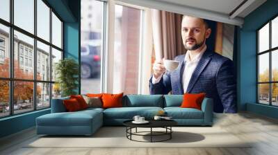 A young man in a business suit is drinking coffee by the window. A young man with a beard in a cafe. Wall mural