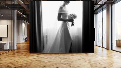  A beautiful bride in a white dress stands near the window and looks at the bouquet. Black and white photo Wall mural