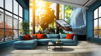 A senior executive standing near a window, holding a tablet displaying complex data reports, while sunlight streams into the room, creating shadows that emphasize the seriousness o Wall mural