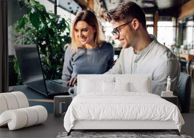 A couple sitting at cafe smiling cheerfully, using a laptop, spending time with pleasure, drinking coffee. Dressed in casual clothes. Wall mural