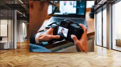 Close-up view of male hands holding professional camera, looks photos, sitting at cafe with laptop. Wall mural