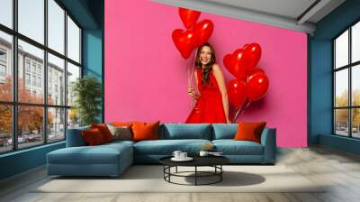 Smiling girl holding air balloons in two hands, posing at camera. St. Valentine's day. Dressed in red dress, on pink background. Wall mural