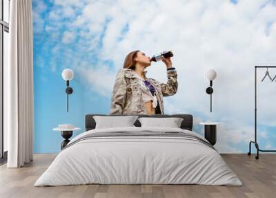 Stylish girl drinking a soda from bottle, standing against the sky background Wall mural