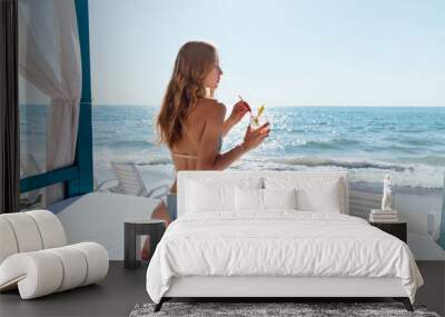 View from back on woman with slim body and long hair, drinking a cocktail, while sitting on a luxury sunbed, near the ocean. Wall mural