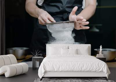 Cropped image of female pastry chef sifting flour for homemade baking, woman in apron holding sieve surrounded by baking ingredients. Wall mural