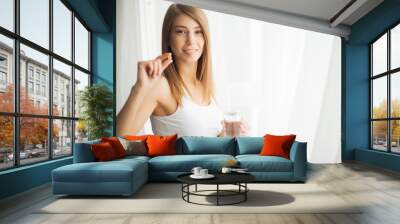 Happy smiling positive woman eating the pill and holding the glass of water in the hand, in her home Wall mural