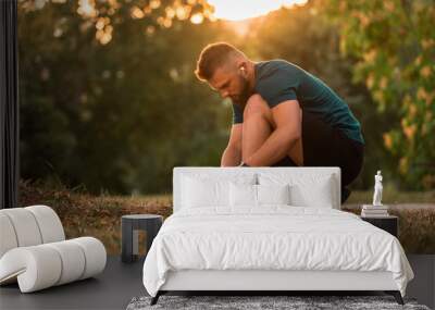 A young athletic man tying running shoes with wireless earphones listening a music for motivation preparing for a run or workout at the park active lifestyle. Wall mural