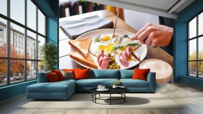 The man eat. Knife and fork in hand. Breakfast on the table.. American style breakfast with fried eggs, sausage, green peas and toast. The concept of a healthy morning. Wall mural