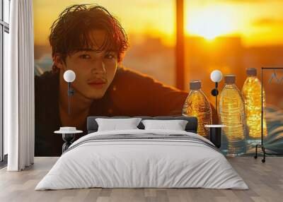 A young man lies on a bed, looking at the camera. There are three bottles of water on the bed with him, and the sunset can be seen in the background through a window. Wall mural