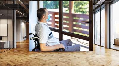 young man wearing face mask sitting in a wheelchair alone looking out the window Wall mural