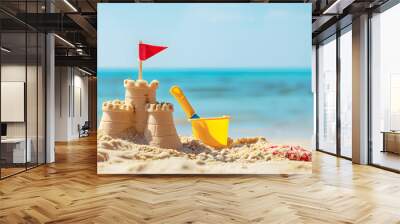 A simple, small sand castle is shown in the foreground with a blue sea background. The sand castle has a flag toy and bucket placed near it on the sunny beach day Wall mural