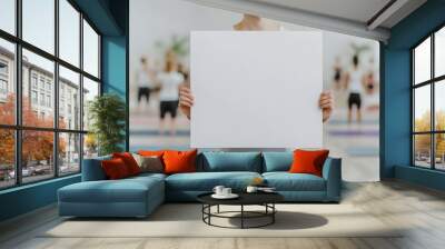 A person holds a blank white sign in a yoga studio, with others practicing yoga in the background, creating a serene and minimalist atmosphere. Wall mural