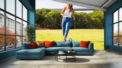 Carefree young woman playing soccer in park. Woman in casual kicking ball on grass. Active lifestyle concept Wall mural