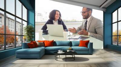 Cheerful coworkers using laptop. Smiling multiethnic businessman and businesswoman sitting at table and working with laptop computer in office. Technology and business concept Wall mural