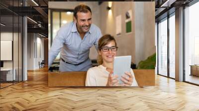 Cheerful woman using pc tablet, her colleague standing behind. Young businesswoman wearing glasses sitting in lobby, male manager standing nearby looking at screen. Wireless technology concept Wall mural